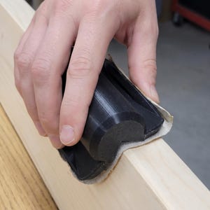 Könnte beinhalten: Ein schwarzer Schleifklotz wird verwendet, um die Kante einer hellen Holzplatte zu glätten. Der Klotz wird von einer Hand gehalten, und das Schleifpapier ist an den Rändern des Klotzes sichtbar. Das Holz ist unbehandelt.