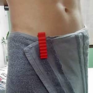 May include: A red plastic towel clip securing a gray towel. The clip has a ridged design. The towel is wrapped around a person's waist.