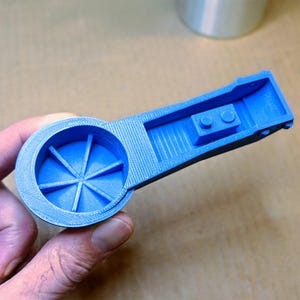 Könnte beinhalten: Hellblaues 3D-gedrucktes Objekt mit einem kreisförmigen Ende mit radialen Linien und einem rechteckigen Griff. Der Griff hat einen kleinen erhöhten Abschnitt mit zwei kreisförmigen Merkmalen. Das Objekt wird in einer Hand gehalten.