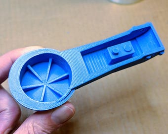 May include: A light blue 3D-printed object with a circular end featuring radial lines and a rectangular handle. The handle has a small raised section with two circular features. The object is held in a person's hand.