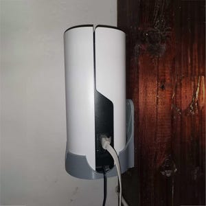 Op de afbeelding: Een wit en zwart cilindrisch apparaat dat met een grijze beugel aan een muur is bevestigd. Het apparaat heeft een gesplitst ontwerp en is aangesloten op kabels. Het apparaat is gemonteerd op een witte muur naast een houten oppervlak.