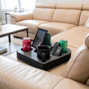 Op de afbeelding: Een zwarte armleuningtray met meerdere compartimenten met een rode blik, een zwarte mok, een groene beker, een tablet, afstandsbedieningen en een groene blik. De tray is ontworpen om drankjes en elektronica op een bank te houden.