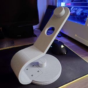 Peut inclure: Un support de casque imprimé en 3D, de couleur gris clair. Il se compose d'une base circulaire, d'un bras de support incurvé avec un trou circulaire et d'un repose-tête. Conçu pour ranger un casque, il mesure en cm.