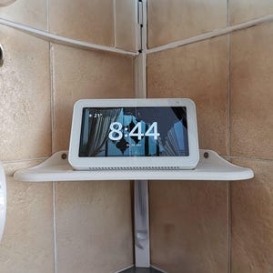Puede incluir: Un dispositivo de pantalla inteligente blanco que muestra la hora 8:44, montado en un estante de esquina blanco. La pantalla muestra una imagen escénica. El dispositivo está en un baño con paredes de azulejos beige.
