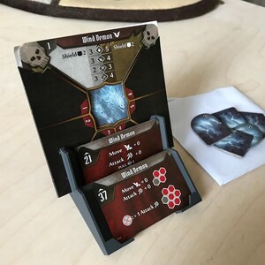 Könnte beinhalten: Ein Brettspiel-Setup mit einer Wind Demon-Karte und einem Kartenhalter. Die Karten sind rot und schwarz mit weißem Text und Grafiken. Zusätzliche Karten mit einem stürmischen Himmelsdesign sind ebenfalls vorhanden.