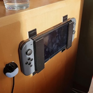 以下が含まれることがあります： 木製の表面に黒いホルダーで取り付けられた、グレーのNintendo Switchコンソール。コンソールは黒いホルダーで壁に取り付けられています。左側には黒と青のプラグが見えます。
