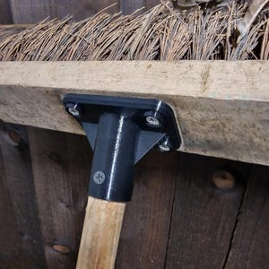 May include: Close-up of a broom head with a black metal bracket and wooden handle. The broom head is made of straw-like material. The bracket is attached to a wooden beam with screws. The handle is made of light-colored wood.