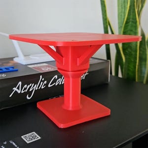 Könnte beinhalten: Ein leuchtend roter, 3D-gedruckter Präsentationsständer mit quadratischer Basis und Tischplatte. Der Ständer hat eine zentrale Säule und abgewinkelte Stützen. Der Text "Acrylic Color" ist auf einer Box sichtbar. Der Ständer steht auf einer schwarzen Oberfläche.