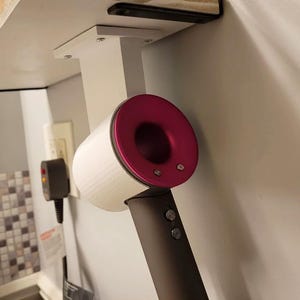 May include: A white and magenta hair dryer is mounted on a white holder attached to a wall. The hair dryer has a dark grey handle with two buttons. The holder is mounted under a shelf.