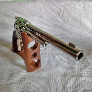 Op de afbeelding: Een glanzende, verchroomde revolver met een donkerhouten handvat rust op een koperkleurige standaard. De standaard heeft drie cirkelvormige uitsparingen. Het pistool staat schuin, waarbij de loop en cilinder worden getoond. De achtergrond is een getextureerde, gebroken witte stof.
