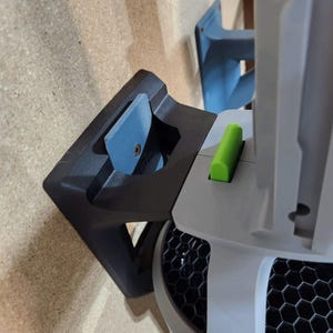 May include: A black and gray mounting bracket attached to a light brown surface. A blue component is secured within the bracket, and a green piece is visible on the adjacent object. The object has a honeycomb pattern.