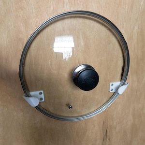 Può includere: Un coperchio per pentola in vetro trasparente con bordo in acciaio inox e pomello nero. Il coperchio ha due clip in plastica bianca ed è progettato per adattarsi a una pentola. Sul pomello è stampata la scritta "Silit".