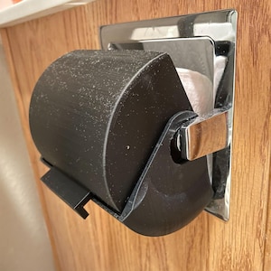 May include: A black toilet roll holder with a chrome metal mounting plate. The holder is mounted on a wooden surface. The toilet paper roll is partially visible inside the holder.
