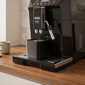 Könnte beinhalten: Eine schwarze De'Longhi Eletta Cappuccino Evo Espressomaschine. Die Maschine hat eine Dampflanze aus Edelstahl und einen Wasserbehälter. Sie steht auf einer Holz-Arbeitsplatte.