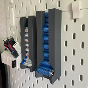 Könnte beinhalten: Drei graue Batterie-Organizer an einer weißen Lochwand befestigt. Die Organizer halten mehrere AA- und AAA-Batterien. Ein weißer Clip und eine Ladestation sind ebenfalls an der Lochwand befestigt.