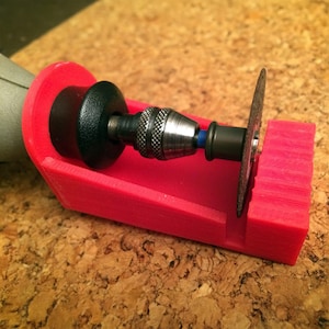 May include: A red 3D-printed rotary tool holder with a cutting disc attached. The tool is partially visible, showing a silver chuck and a black and blue component. The holder is angled and sits on a brown surface.