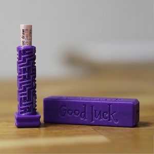 Puede incluir: Un soporte morado para boletos de lotería impreso en 3D y una caja rectangular con "Good Luck" en relieve. El soporte tiene un diseño de laberinto y contiene un pequeño boleto con texto. La caja también es morada.