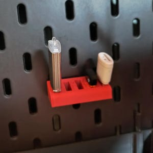 Puede incluir: Un organizador rojo impreso en 3D con múltiples ranuras, que sostiene un bolígrafo vape plateado, un dispositivo negro y un bloque de carga blanco. El organizador está montado en una panel perforado negro con agujeros ovalados.