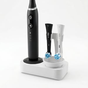 Puede incluir: Un cepillo de dientes eléctrico negro con una base de carga blanca. Dos cabezales de cepillo de dientes, uno negro y otro blanco, están en la base. Los cabezales del cepillo de dientes tienen cerdas azules. La base tiene un cuenco pequeño.