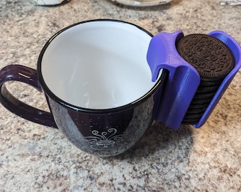 May include: A purple mug with a white interior and a purple cookie holder attached to the rim, holding a stack of chocolate sandwich cookies. The mug has a decorative design on the side. The cookie holder is made of plastic.