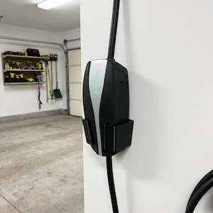 May include: A black and silver electric vehicle charger mounted on a white wall. The charger has a sleek design with a curved silver accent and a series of green indicator lights. A black charging cable extends downwards.