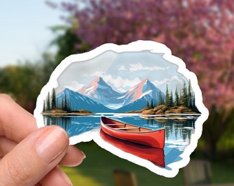 May include: A sticker featuring a scenic landscape with a red canoe on a lake, mountains, and trees. The sticker is held in a hand against a blurred outdoor background with a table and chairs.