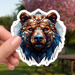 May include: A sticker featuring a geometric design of a bear's head. The bear has a brown and blue color scheme with a blue nose and is held in a hand against a blurred outdoor background.