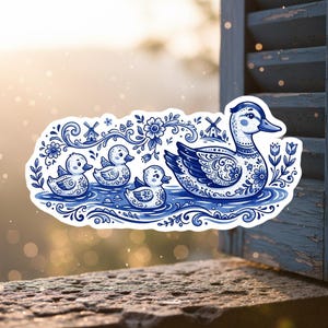 May include: A blue and white sticker featuring a mother duck and three ducklings swimming in water. The design incorporates floral and windmill motifs, with intricate patterns on the ducks. The sticker has a decorative border.