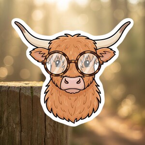 May include: A sticker featuring a cartoon illustration of a Highland cow wearing round, tortoiseshell glasses. The cow has light brown fur, a pink nose, and large curved horns. The sticker is outlined in white.