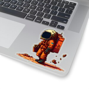 May include: A sticker featuring a pixelated astronaut in an orange spacesuit, skiing on a pixelated trail. The astronaut has a black visor and a white helmet. The sticker is on a laptop.