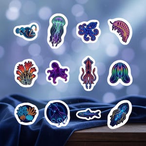 May include: A collection of colorful ocean-themed stickers featuring various marine life. Designs include a jellyfish, octopus, squid, coral, and other sea creatures. The stickers have a white border and are set against a blurred blue background.