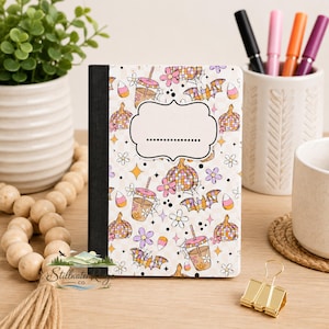 May include: A notebook with a black spine and a white cover featuring a repeating pattern of orange and purple pumpkins, bats, iced coffee cups, candy corn, and flowers. A central white label with a dotted line is printed on the front cover.