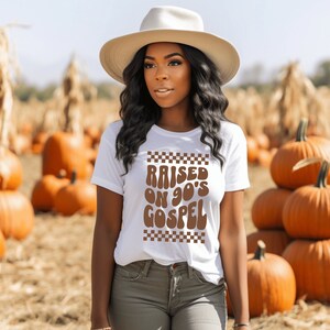 Op de afbeelding: Wit T-shirt met bruine tekst die "RAISED ON 90'S GOSPEL" luidt, met een geruit patroon boven en onder de tekst. Een persoon draagt het shirt in een veld met pompoenen.