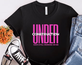 May include: Black t-shirt with the text "UNDER CONSTRUCTION" in pink letters and the text "GOD'S STILL WORKING ON ME" in white letters.