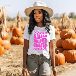 Op de afbeelding: Een wit T-shirt met een roze afbeelding waarop in een retro-lettertype "RAISED ON 90'S GOSPEL" staat. De afbeelding is omgeven door een geruite rand.