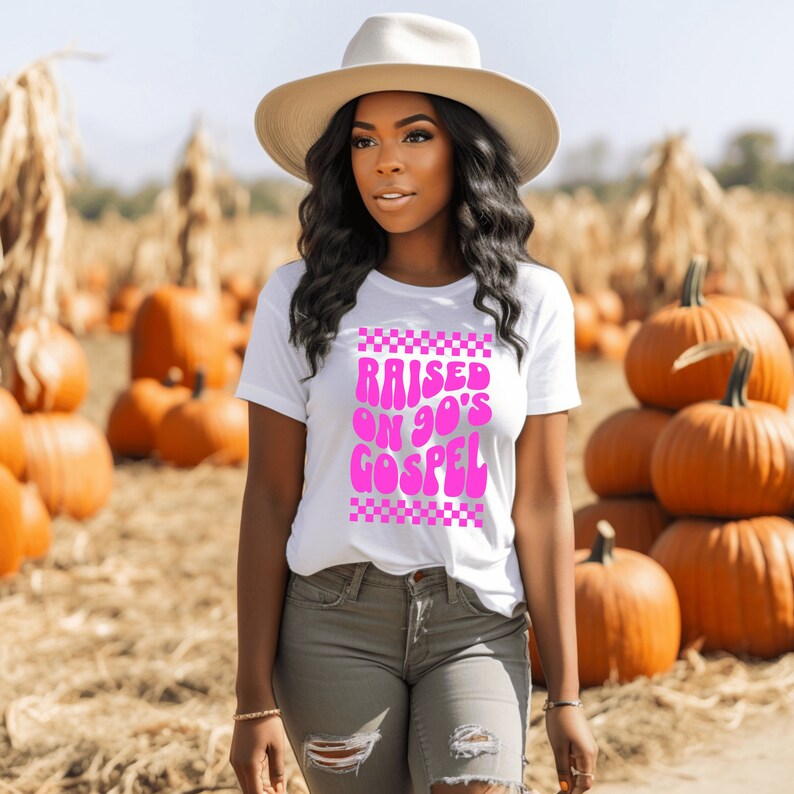 Op de afbeelding: Een wit T-shirt met een roze afbeelding waarop in een retro-lettertype "RAISED ON 90'S GOSPEL" staat. De afbeelding is omgeven door een geruite rand.