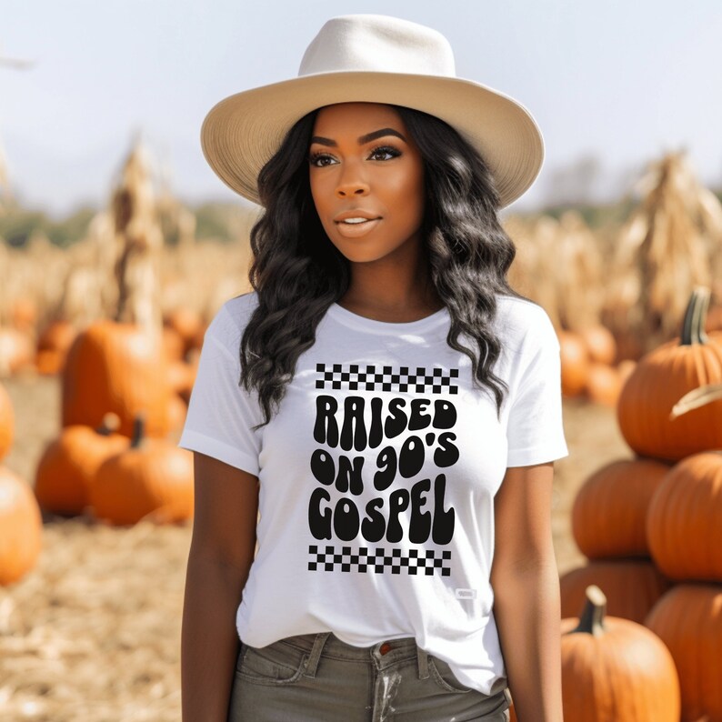 Op de afbeelding: Een wit T-shirt met een zwarte afbeelding waarop "Raised on 90's Gospel" staat in een retro lettertype. De afbeelding is omgeven door een zwart-witte geruite rand.
