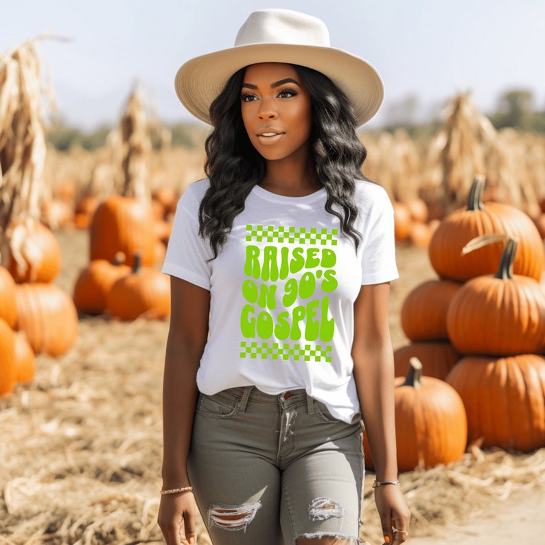 Op de afbeelding: Een wit T-shirt met een groene afbeelding waarop in een retro-lettertype "Raised on 90's Gospel" staat. De afbeelding is omgeven door een geruite rand. Het model draagt het shirt en staat in een veld met pompoenen.