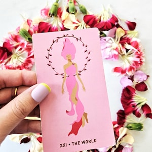 May include: A pink tarot card with the words "XXI • THE WORLD" at the bottom. The card features an illustration of a woman with pink hair and a pink dress, surrounded by a wreath. The card is surrounded by red and white flower petals.