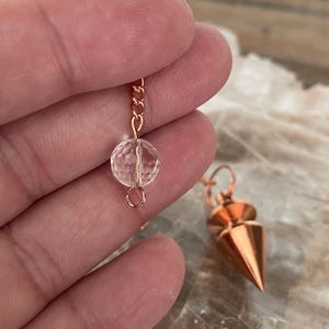 May include: A rose gold pendulum with a clear crystal sphere and a pointed cone-shaped weight. The pendulum is suspended by a delicate chain. The crystal sphere is approximately 0.5 inches in diameter.
