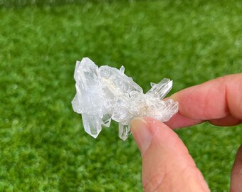 May include: A cluster of clear quartz crystals, held between fingers, against a blurred green background. The crystals are translucent and have a natural, jagged formation. The quartz appears to be raw and unpolished.