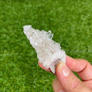 Può includere: Un gruppo di cristalli di quarzo trasparente, tenuto contro uno sfondo verde. I cristalli di quarzo sono trasparenti e hanno una struttura cristallina. La base del gruppo è di colore bianco sporco.