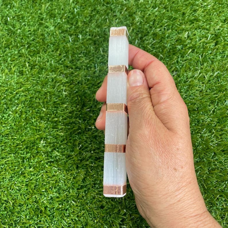 May include: A white selenite wand with copper wire wrapped around it. The wand is held in a hand against a green grassy background.