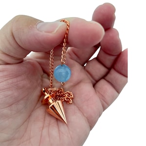 Może przedstawiać: Różowe złoto wahadło ze spiczastą końcówką i jasnoniebieskim kryształowym koralikiem. Wahadło wisi na pasującym łańcuszku. Kryształowy koralik jest okrągły i gładki.