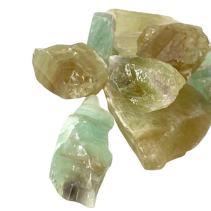 May include: A collection of raw, uncut fluorite crystals in shades of green and yellow. The natural stones have a rough, textured surface and varying shapes. The crystals are displayed against a white background, highlighting their natural colors and textures.
