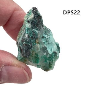 May include: A rough-hewn, natural mineral specimen with a mix of teal, dark green, and black hues. The surface has a textured appearance, with the text "DPS22" in the upper right corner. The mineral is held in a person's hand.