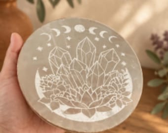 May include: A round, light gray stone coaster featuring a white engraved design of a crystal cluster surrounded by succulents. The top edge displays a lunar cycle phase pattern with small stars scattered throughout the central illustration.