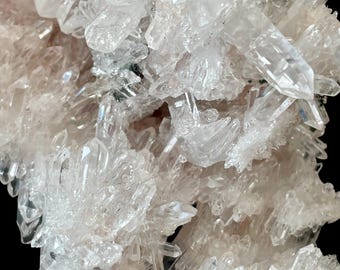 May include: Close-up of a cluster of clear quartz crystals. The crystals are translucent with sharp, pointed terminations, and are clustered together in a dense formation. The overall color is a pale, off-white with hints of pink.