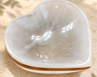 May include: A heart-shaped bowl carved from translucent white selenite crystal. The interior surface is smooth and concave, while the exterior features a distinct horizontal band of warm brown wood inlay encircling the base of the white stone.
