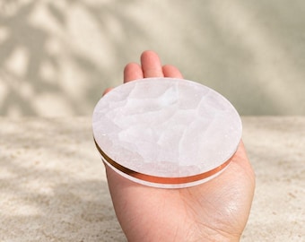 May include: A circular coaster made of translucent, pale pink rose quartz with a textured, uneven top surface. The side edge of the coaster features a smooth, polished copper-colored metallic band that encircles the entire circumference.